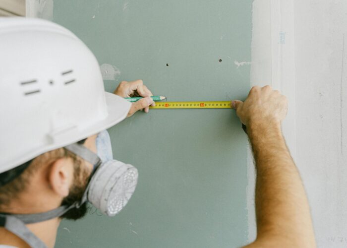 A construction worker precisely measuring drywall, wearing a helmet and mask for safety.